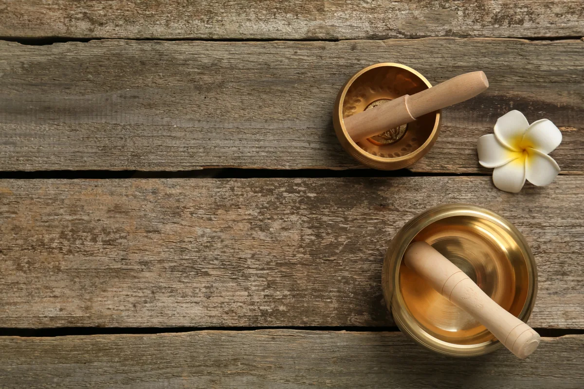 Zwei goldfarbene Klangschalen mit hölzernen Klöppeln liegen auf einem alten Holztisch, daneben eine weiße Blüte.