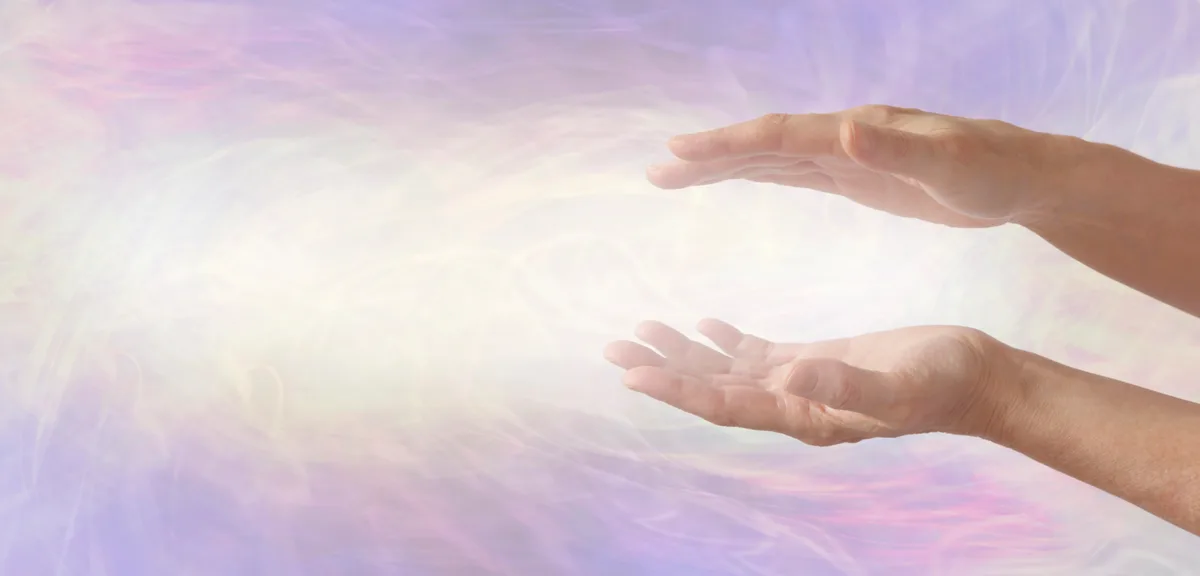 Zwei geöffnete Hände schweben übereinander vor hellem, zart violett und rosa schimmerndem Licht.